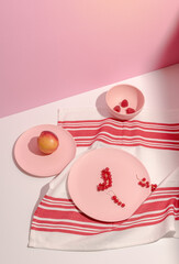 Trending colors. Plates with fruits and berries on tablecloth with red and white grid on pink background. Isometric view minimal still life. Minimal summer composition. Long harsh shadows. Copy Space