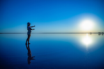 Lovely happy young girl walks and poses in a magical pose along the mirror pink salt lake enjoying the warm evening sun, looking at the fiery sunset and her reflection
