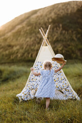 cute little girl is having fun near wigwam in a summer field on sunset. Young family spending time together on vacation, outdoors. The concept of summer holiday. © Andriy Medvediuk