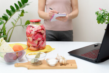 Obraz na płótnie Canvas Cooking at home a woman watches online video recipes on a laptop and cooks in the kitchen at home writes in a notebook. Pickled pink cabbage fermentative with beets and carrots in brine in a jar