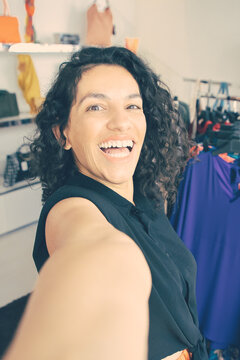 Cheerful excited Latin woman taking selfie near rack with dresses in fashion shop, looking at camera, smiling and laughing. Boutique customer or shop assistant concept