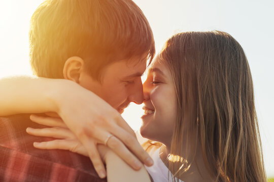 Cropped close-up side view of happy blond woman and handsome man hugging tenderly outdoors on sunset background. 
