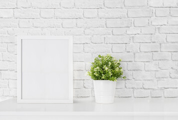 White frame mockup with green plant on a white table against white brick wall. Empty poster frame mockup for design. Copy space