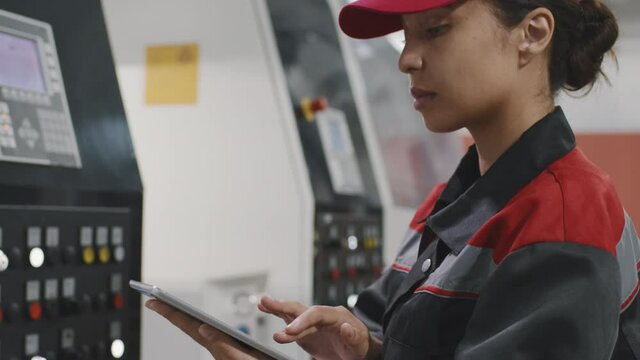 Tilting-down slow motion close-up of female mixed race factory worker using digital tablet while setting parameters for modern machine equipment