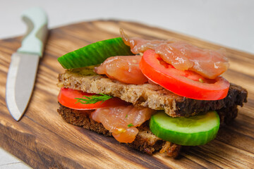 Trout sandwich with wholegrain bread, cucumber, herbs and tomatoes laying on a rustic board with kitchen knife