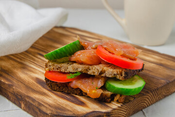 Red fish sandwich with wholegrain bread, cucumber, herbs and tomatoes laying on a wooden board