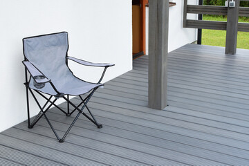 Part of white residential house with dark gray or anthracite wpc composite material terrace and empty chair on deck with wooden railings outdoors.
