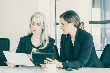 Focused professionals analyzing reports together. Two businesswomen sitting together, reading documents, using tablet and talking. Teamwork concept