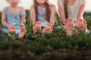 Three sisters in white sundresses are sitting on green meadow. Girls play in blooming field of purple sage. Show their feet heels Happy family love. Outdoor walks, happy barefoot childhood. Copy space