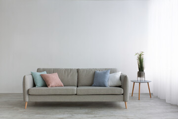 Gray sofa with pillows, table with plants and white curtains on window, on light wall background in living room