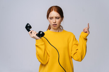 Desperate young woman in stylish yellow sweater holding handset receiver of retro phone and looking on him against white background. Pretty redhead lady emotionally showing facial expressions.