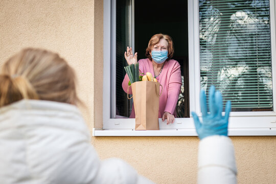 Social distancing due coronavirus covid-19 pandemic lockdown. Senior woman with face mask waving from window to her adult daughter she delivered groceries