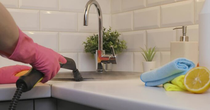 Housewife in rubber gloves cleaning black kitchen sink with a hot steam cleaner and blurred cleaning products, citron and sponges on kitchen countertop.