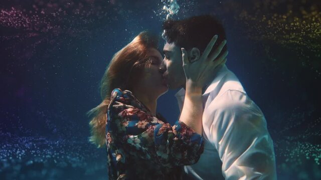 Cinematic shot of young carefree romantic loving couple is kissing with passion in blu underwater with colorful water splash. Concept of love, relationship, freedom, sensuality, fantasy.
