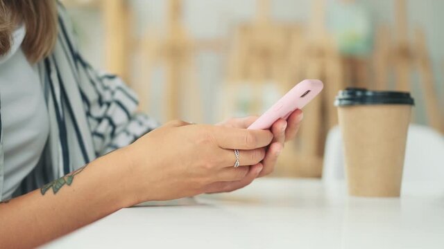 A cropped close-up side view of a young woman is scrolling on her smartphone while sitting at the table