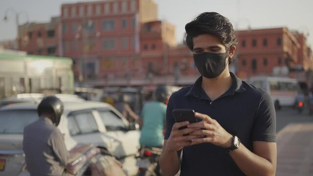 close view shot of young Indian male with protective mask on face using a mobile phone to text a message while walking on a footpath along with busy road traffic around