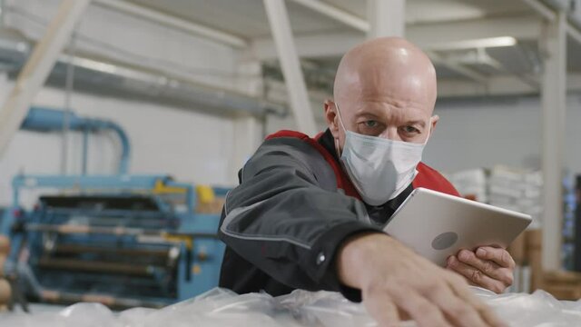 Medium slowmo close-up of caucasian factory workman in protective mask checking quality of produced polyethylene making notes on digital tablet