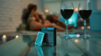 Sexual adventure. Two glasses of red wine and condoms on the table with young half naked man and woman kissing and cuddling on the bed before making love