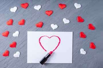 Fototapeta premium Lipstick draws a heart shape on white paper on a gray background with white and red hearts, copy space, flat lay, top view, flat lay. Valentines day background