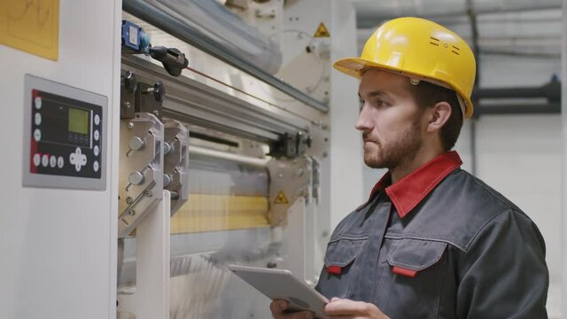 Medium slowmo closeup of male engineer in coverall and helmet working on digital tablet setting parameters for machine equipment producing polyethylene film