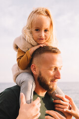 Daughter sits on dad's shoulders. Walking next to the sea. Baby girl with blonde hair, in a warm fur vest. Spring time, sun is shining. Travel abroad. Happy young family. 
