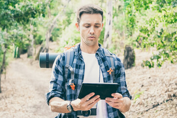 Naklejka na ściany i meble Young backpacker hiking, holding tablet and looking at map. Caucasian attractive traveler walking on road in woods. Trees on background. Backpacking tourism, adventure and summer vacation concept
