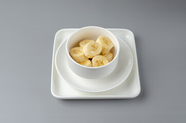Slices of bananas in a white plate on a gray background. Trending color Pantone of year 2021 Illuminating and Ultimate gray. Top view minimal still life.