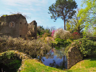 Fototapeta na wymiar Garden of Ninfa or Giardino di Ninfa with medieval ruins old river bridge and flowering exotic plants of various colors. Amazing park of Caetani family during spring. Beautiful landscape with blue sky