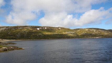Fototapeta premium The sun's rays illuminate the hills on the opposite shore of the Barents Sea bay. View of the coast. Northern summer landscape. Snow in the hills in summer.