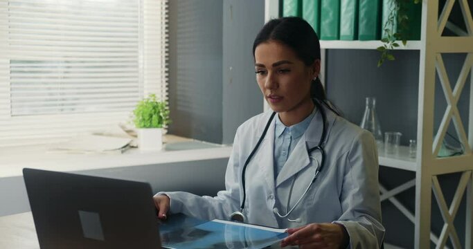 Serious female doctor pulmonologist talking to sick patient online via video call on laptop, holding lungs x-ray and comments diagnosis.