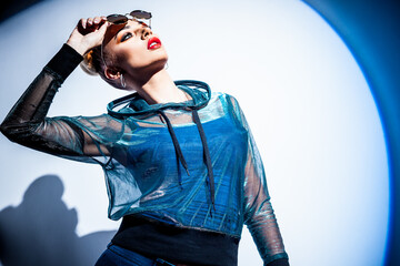 Fashion concept. Portrait of a beautiful woman in jacket with hood and sunglasses in bright mixed contrast light. Female model dances in studio. Professional makeup and colorful background