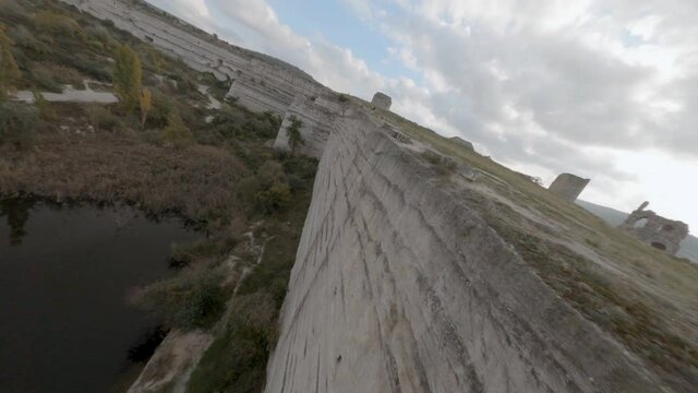 Drone freestyle flying along flat white cliff edge and ancient fortress. Aerial view antique wreckage fortress and lake. Historical architecture on mountain plateau from bird above. Fpv sport copter.