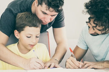 Gay parents helping focused boy with school home task, sitting at table and writing in papers together. Family and parenthood concept