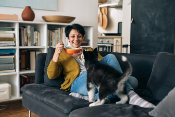 woman sitting on sofa in her living room and having snack, her dog sitting beside her