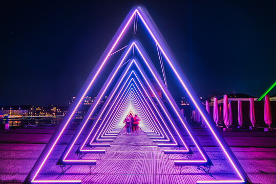 Fototapeta Purple coloured gate of light or purple light tunnel installation made of triangular neon and led lights at night as blurred people walk through the bright deep passage that resembles a human trachea