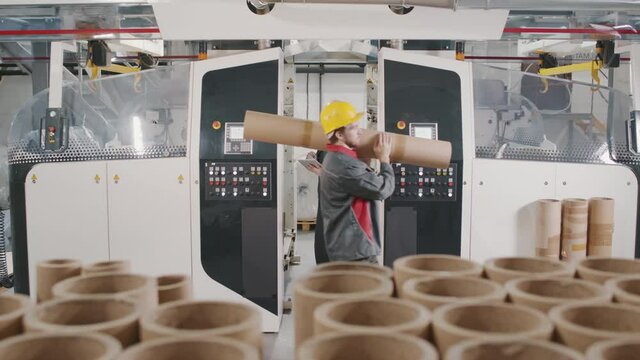 Slow-motion medium footage of workmen in coverall uniform working on production of spiral cardboard tubes for winding up polyethylene