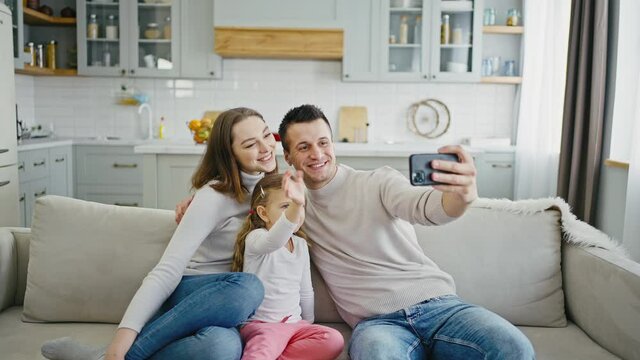 Mother, father and little daughter sitting together on couch and smiling to smartphone camera, making selfie