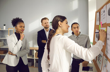 Professional team of multiracial business people brainstorming in office. Young woman writing idea on sticky note pinned to bulletin board in group meeting with multiethnic coworkers. Teamwork concept