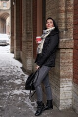 a beautiful girl with shoulder-length dark hair stands in winter clothes, leaning her back on a stone wall and holding a red disposable cup in her hands