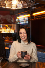 a beautiful girl with graceful features and dark shoulder-length hair sits in a cafe, laughs and holds a mug in her hands