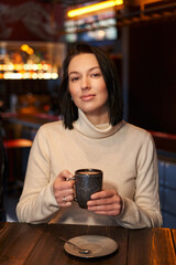 a beautiful girl with shoulder-length dark hair holds a mug in her hands and sits in a cafe