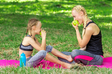 Mother with daughter in sportswear eats apples sitting on a mat in the summer park