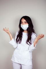 young asian woman doctor in white uniform gesturing positive on gray background, wearing a mask,...