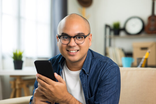 Young asian man wearing glasses sitting on sofa in living room and using smartphone while work from home.