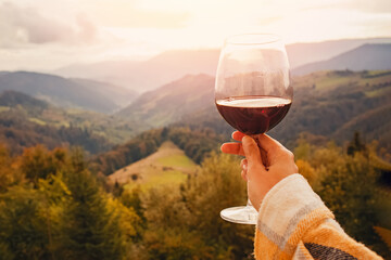 Woman's hand holding a glass of red wine with a mountain view. - obrazy, fototapety, plakaty