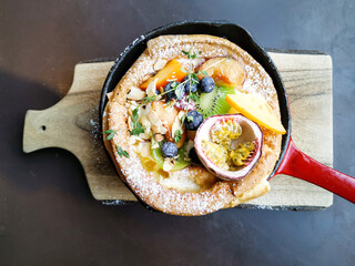 Delicious homemade puff pancake, cooked with vanilla yogurt and fresh fruits, served in vintage red pan on a wooden plate for breakfast