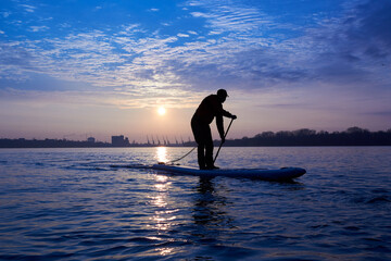Silhouette of active man rowing with paddle on SUP at winter river at sunrise