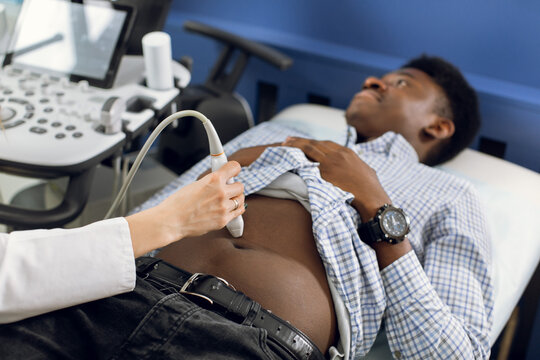Cropped close up image of ultrasound scanner device in the hand of a professional female doctor examining young African man patient, doing abdominal ultrasound scanning sonography