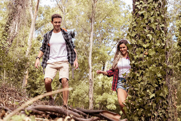 Naklejka na ściany i meble Happy couple traveling together and hiking in forest. Two Caucasian backpackers walking through woods, chatting, smiling and enjoying nature together. Tourism, adventure and summer vacation concept