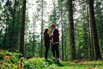Stylish couple of men and women in red shirts on a hike stopped to rest in a coniferous forest, standing on the moss against the backdrop of a beautiful landscape and talking.